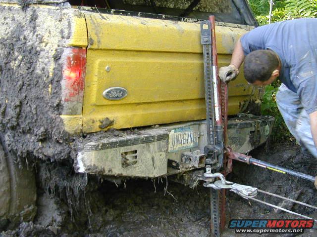 Yellow Ford truck stuck in mud, being lifted by a Hi-Lift jack. License plate UN4GVNG visible