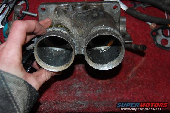 Hand holding a dirty dual throttle body part. Other engine parts and a gasket are visible in the background