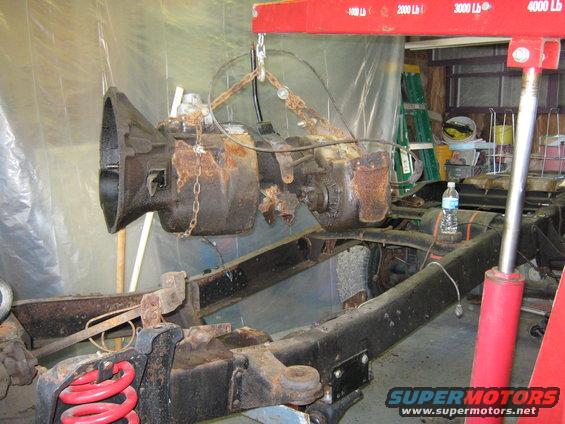 Rusty transmission being lifted by a red engine hoist above a truck chassis with red springs