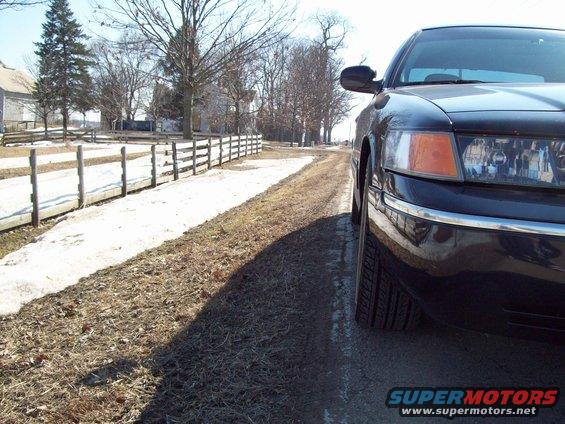 Front corner of a black sedan parked on a dirt road next to a wooden fence. Snow is on the ground