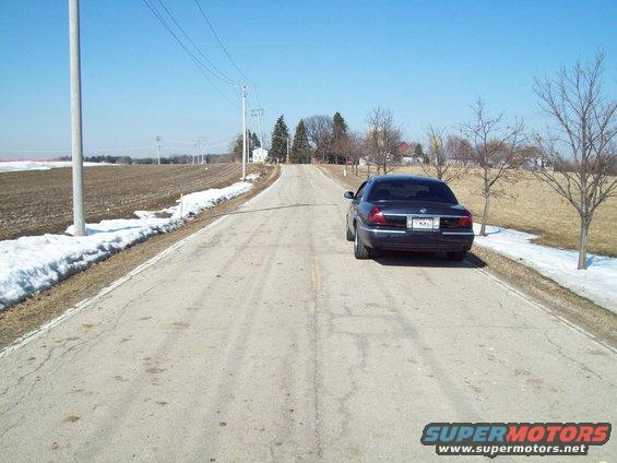 dark sedan parked on side of a paved rural road, snow on the shoulder