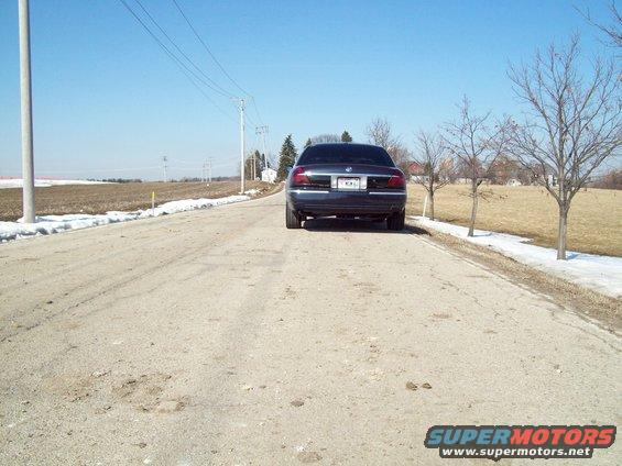Dark sedan from rear on a dirt road. License plate is visible