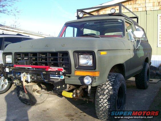 Olive green Ford Bronco with roof rack, winch, and large tires. Another vehicle is partially visible to the left