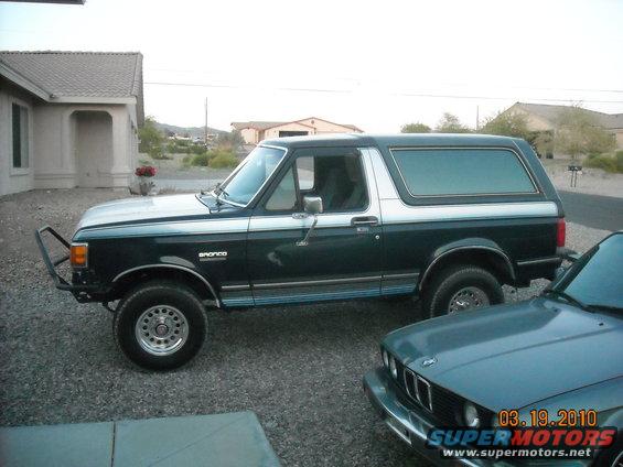 1988 Ford Bronco, dark green with silver top, aftermarket front bumper. Another car is partially visible in the foreground