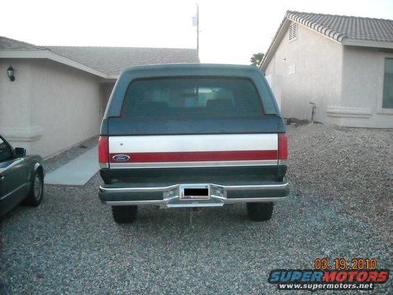 Rear view of a dark blue and silver Ford Bronco with a red stripe. It's parked in a gravel driveway in front of a house