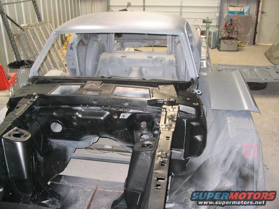 Car body in primer, front view, undergoing restoration. Black engine bay visible