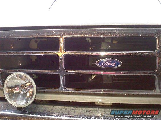Close-up of a Ford emblem on a chrome grille. A round headlight is visible on the left