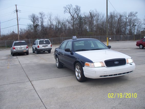 Front view of a dark blue and white Ford Crown Victoria taxi with a "929" sign on its roof. Two SUVs are parked behind it