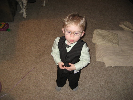 Child wearing a light green shirt, dark pinstripe vest, black pants, and glasses stands on carpet