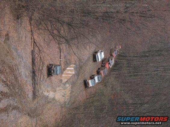 Multiple off-road vehicles driving in a line on a muddy, wooded trail