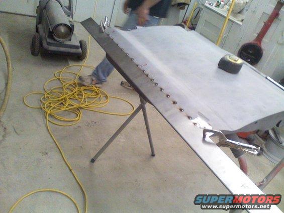 car door panel on sawhorses with welding spots, tape measure on top, yellow cord on floor, heater in background