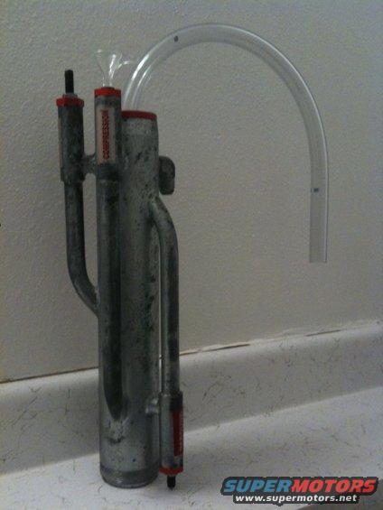 Metal automotive AC accumulator with red caps and a clear hose attached. Text 'COMPRESSION' is visible