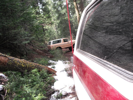 Tan SUV stuck on a muddy, snowy trail with a fallen tree nearby. Another vehicle is in the foreground