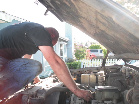 Man in jeans and black t-shirt working on a car engine under an open hood