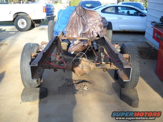 Truck chassis on blocks, covered load on top. White pickup and white coupe in background