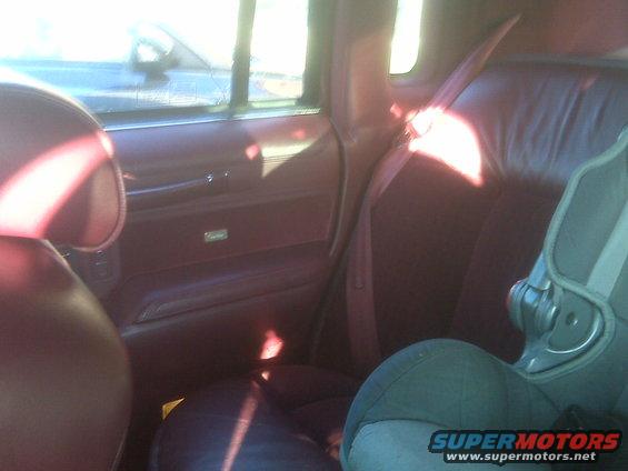 Car interior showing maroon seats and door panel, with a child seat installed on the rear seat. A watermark reads SUPERMOTORS