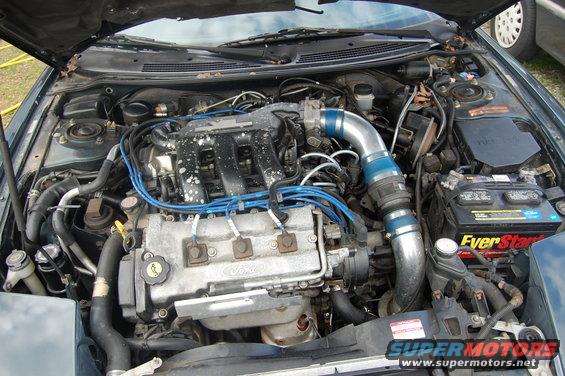 Engine bay with Ford V6 engine, blue spark plug wires, and aftermarket intake. Battery is visible
