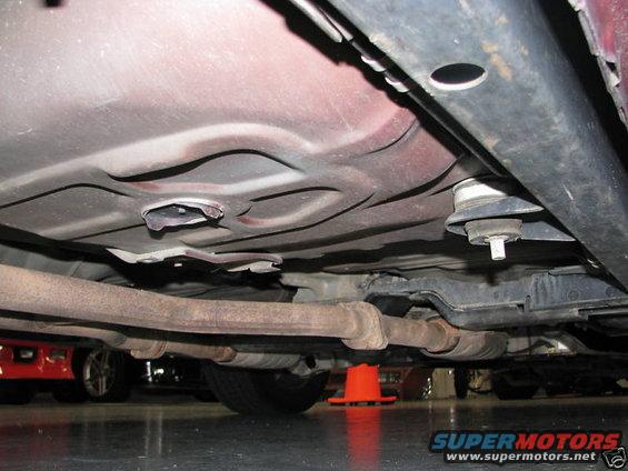 Underside of a car showing exhaust pipes, frame, and suspension components. Another car is visible in the background