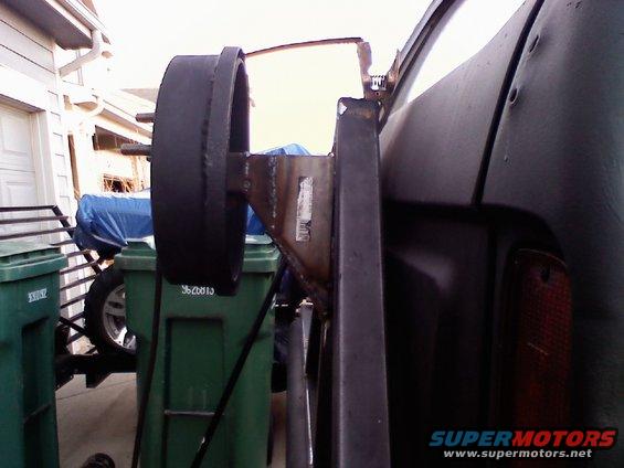 close-up of a custom spare tire carrier mount welded to a vehicle's rear quarter panel, with trash cans and a tarp-covered object in the background