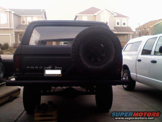Rear view of a black Ford Bronco with a spare tire on the tailgate and a custom bumper. A white pickup truck is parked to the right