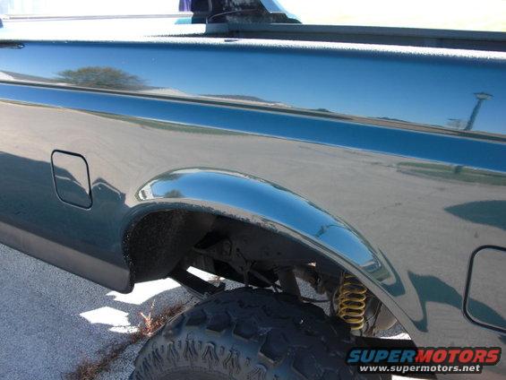 Dark green pickup truck rear fender with chrome trim, visible yellow shock absorber and large tire. Watermark reads SUPERMOTORS