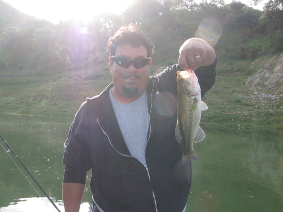 Man in sunglasses and a zip-up jacket holds up a largemouth bass he caught while fishing on a lake