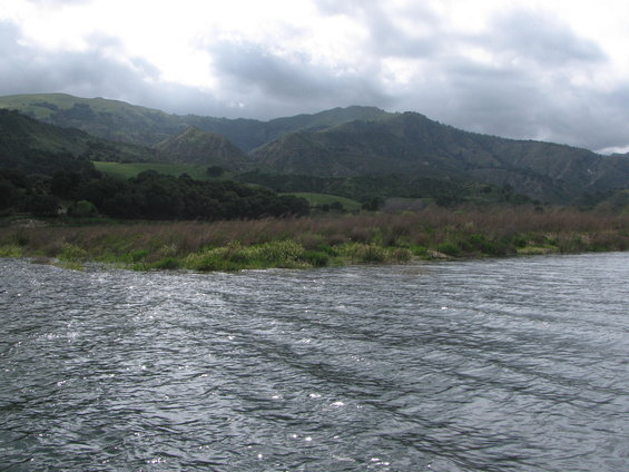 Water ripples in foreground with grassy bank and rolling hills under a cloudy sky