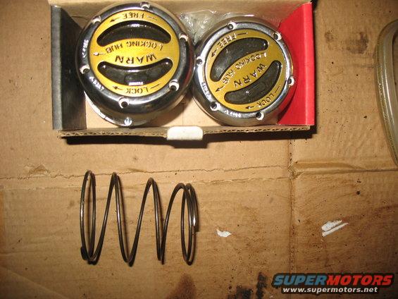 Two chrome and yellow Warn locking hubs in a box with a metal spring on cardboard
