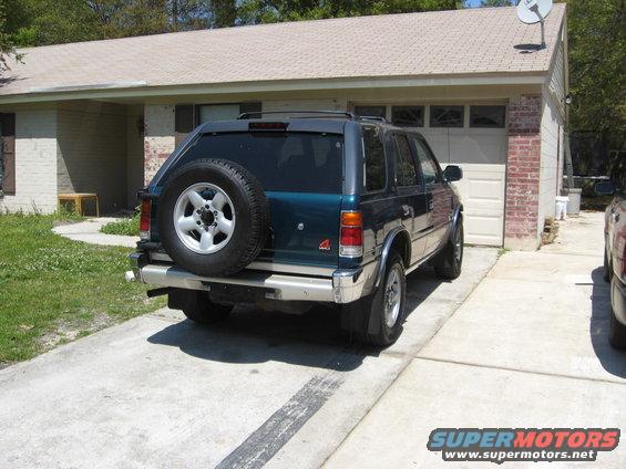 Teal SUV parked in driveway, rear view. Spare tire on back, chrome bumper, mud flaps. House and garage in background