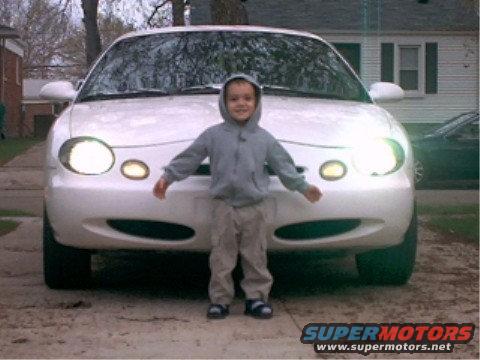 White 1997 Ford Taurus with headlights on, viewed from the front with a child standing before it