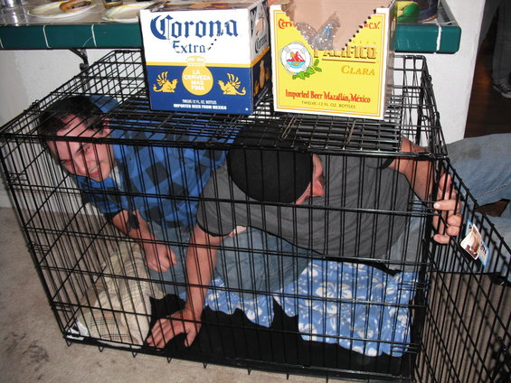 Two men crammed inside a dog crate with Corona and Pacifico beer boxes on top