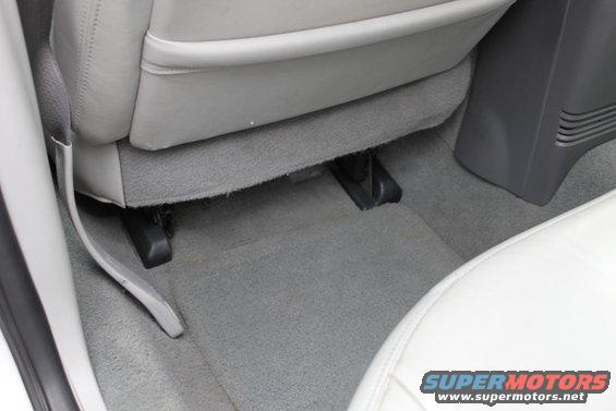 Car interior floor with gray carpet and light gray seats. Shows seat rails and part of a center console