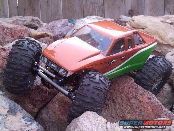 RC rock crawler truck with oversized tires climbing over rocks. Orange and green paint scheme