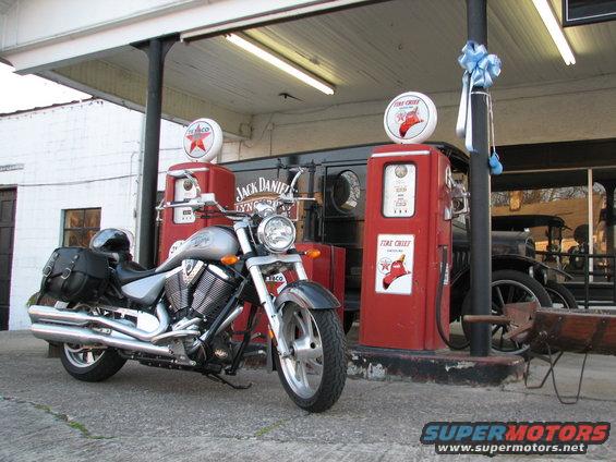 Silver motorcycle parked at vintage gas station with two red gas pumps and a classic car in the background
