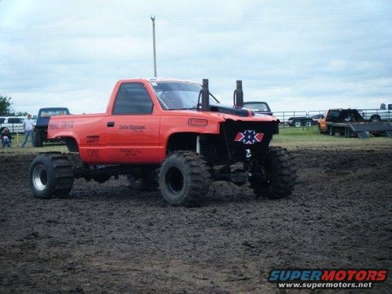 Orange pickup truck with large tires and exhaust stacks in mud. Other vehicles and people in background