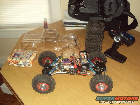 RC rock crawler chassis, clear pickup truck body, stack of tires, remote control, and parts box on a wooden table