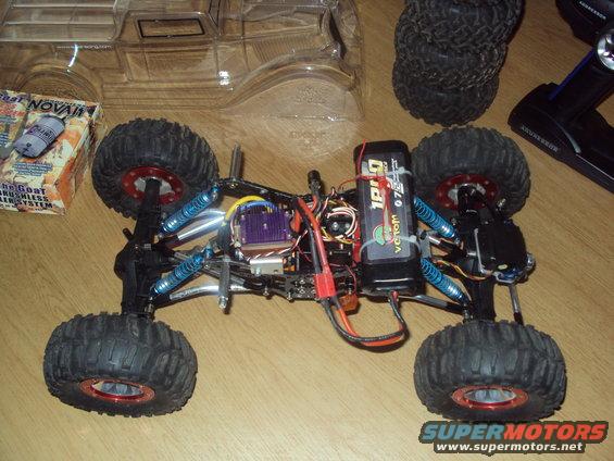 RC rock crawler chassis with battery, remote, and spare tires. Box for a "The Goat" brushless system is nearby