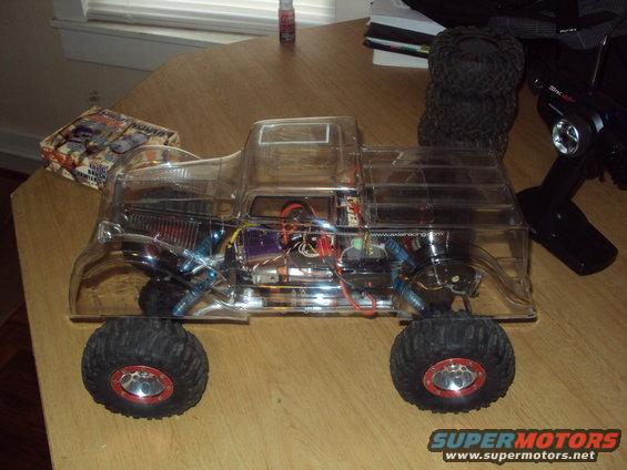 RC truck with clear body, showing internal components and large tires. Remote control transmitter is visible to the right