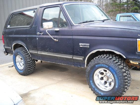 Dark blue Ford Bronco, side view, lifted with off-road tires. Visible "Bronco" badge