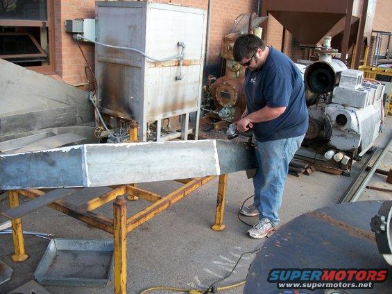 Man in blue shirt and jeans grinding a metal beam on a yellow stand, with industrial equipment in the background