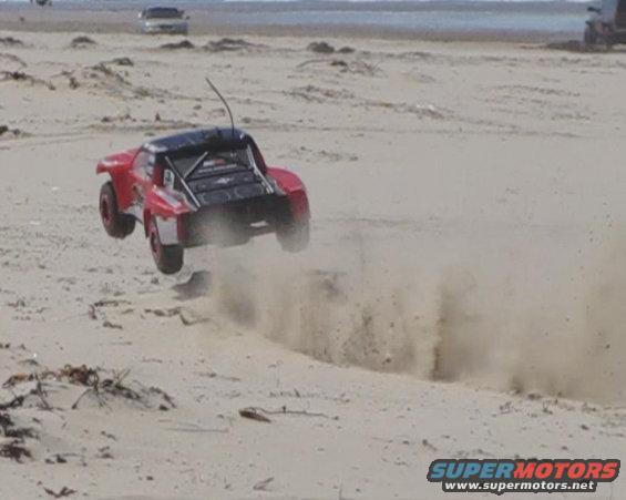 Red remote control truck airborne over sand, kicking up dust. Two other vehicles visible in the distance
