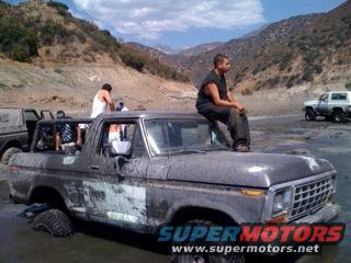 Muddy 1978 Ford Bronco stuck in water with people on top and in the back. Another Bronco is visible in the background