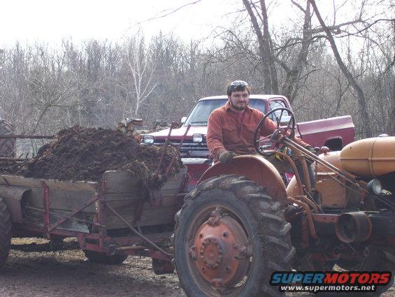 Tractor with a man driving, pulling a trailer full of manure. Red pickup truck behind it