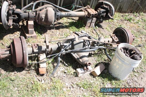 Two vehicle rear axles are sitting on the ground, supported by bricks. A white bucket filled with parts is nearby