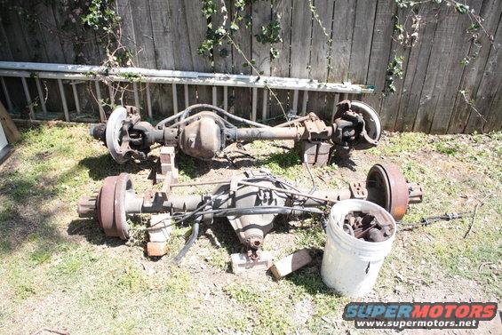 Two rusty truck rear axles are displayed outdoors on grass in front of a wooden fence. A white bucket filled with metal parts sits to the right of the lower axle