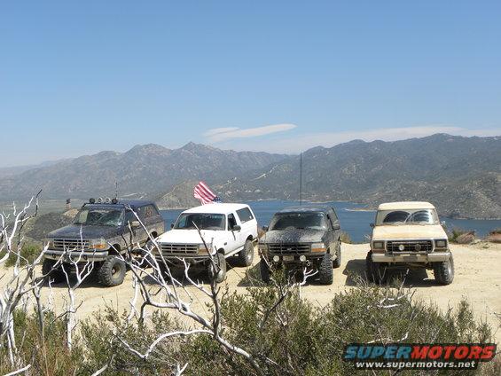 Four SUVs parked on a dirt road with a lake and mountains in the background. One has an American flag