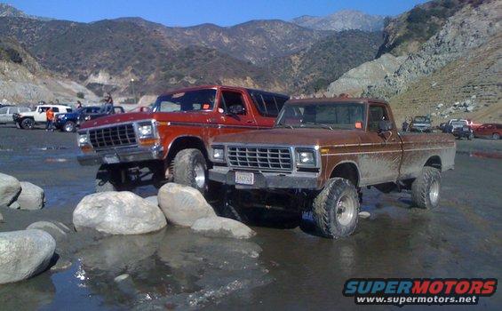 Two lifted Ford trucks, a red Bronco and a brown pickup, parked in shallow water with mountains behind them