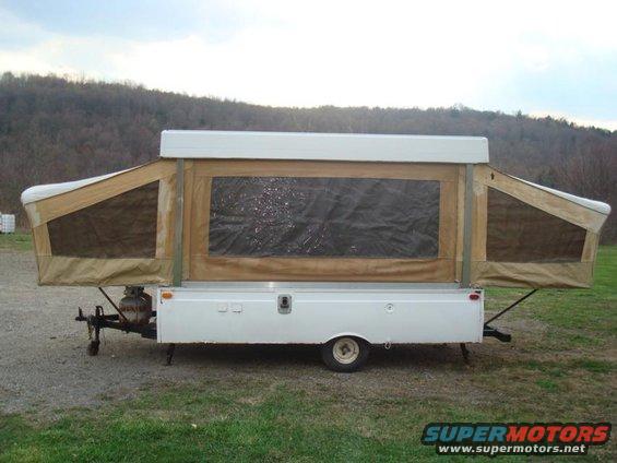 Pop-up camper trailer with tan canvas sides and a white roof, parked on a gravel surface with a wooded hill in the background