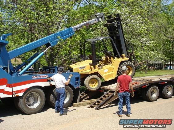 Tow truck with blue boom lifting yellow forklift onto a flatbed trailer. Two men are present