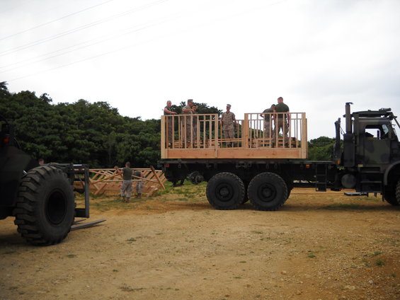 Military truck with flatbed carrying a wooden structure, with soldiers on top. A forklift is to the left